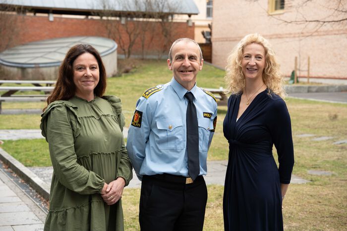 Tre personer: en mann og to kvinner. Mannen står i midten og har på seg politiuniform, kvinnene er kledd i sivile klær. Alle tre smiler inn i kamera. De står foran en gressplen