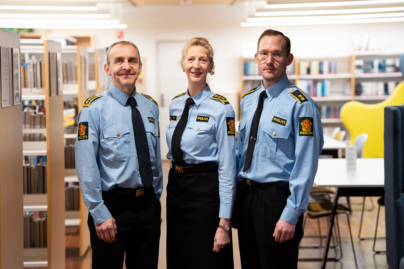 Tre personer i politiuniform foran hyller med bøker. To menn og en kvinne.