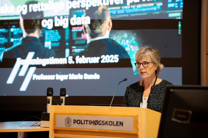 En kvinne med lyst hår og briller står ved et podium det står "Politihøgskolen" på foran en skjerm med bilde av to menn foran mange dataskjermer.