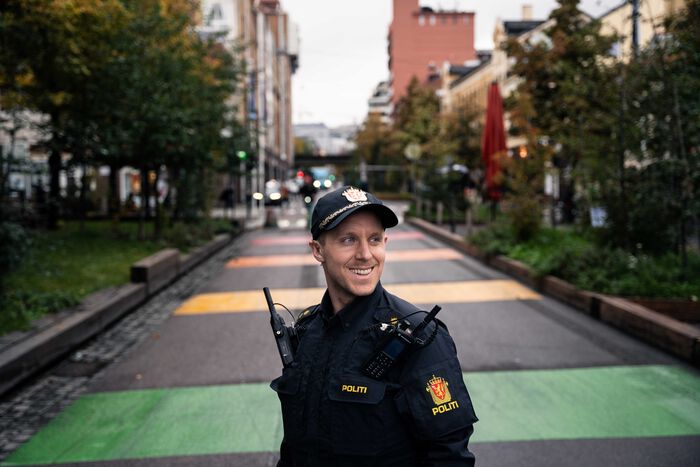 Bilde av en smilende politimann i svart uniform. Han står foran en gate som er dekket i striper i forskjellige farger.