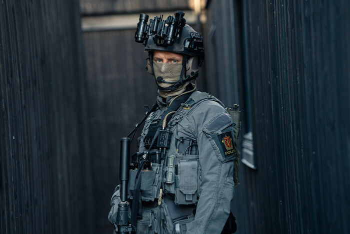 Mann i kjeledress med politilogo på armen. Han har på maske og hjelm, og bærer våpen