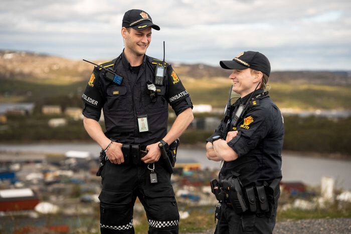 To politimenn i uniform står og snakker sammen. I bakgrunnen ser vi bebyggelse.