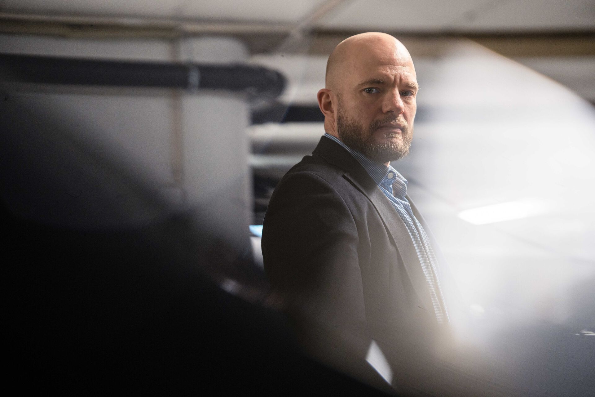 Mann i mørk dress står foran en bil og ser alvorlig inn i kamera