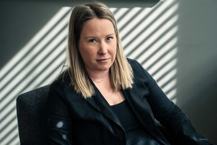 Blond kvinne i svart dress ser rett inn i kameraet uten å smile. Hun sitter foran en lys vegg med skygge fra persienner.